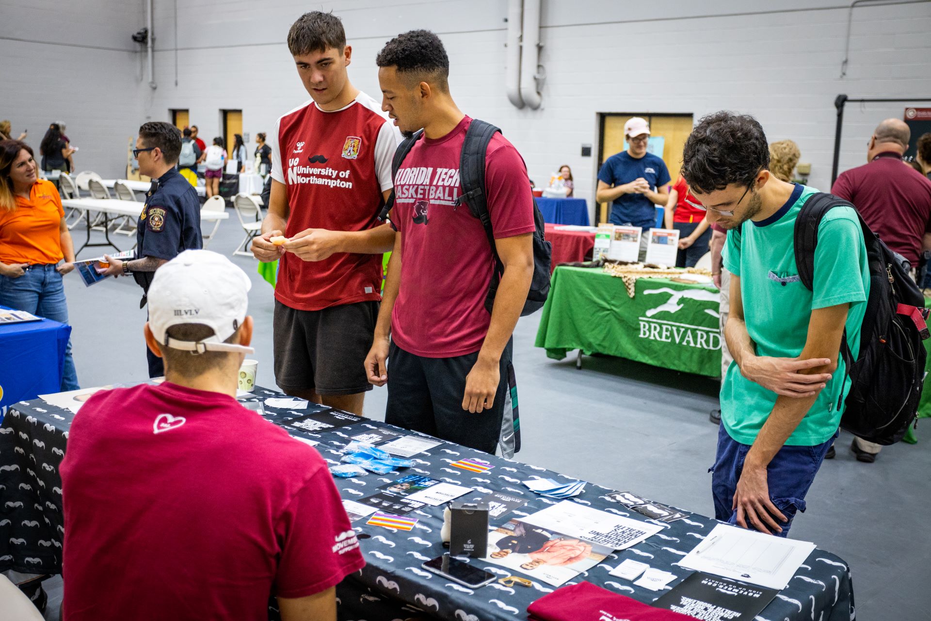 Students interacting at a booth during an event at Florida Tech. Two students in red shirts and one in a green shirt are examining materials on the table. Other attendees and booths are visible in the background